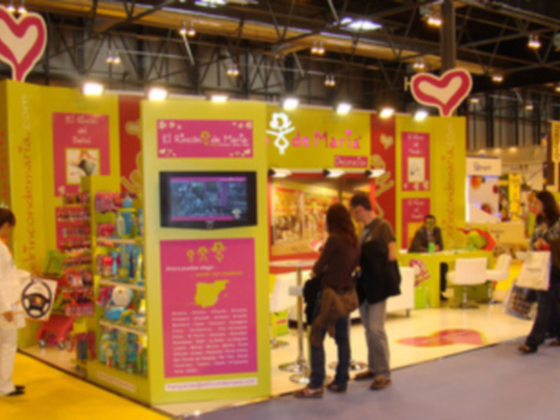 Exposición de productos infantiles en una feria. Stand con estanterías y carteles promocionales. Público interactuando en el stand.