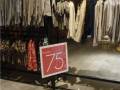 En una tienda de ropa, colgadas en perchas se pueden ver prendas de vestir. En el centro hay un cartel con la oferta "hasta 75%".