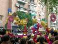 Un desfile de carnaval con figuras inflables de los Simpson, incluyendo a Bart y Homer. La escena se desarrolla en un entorno urbano con edificios y árboles al fondo.