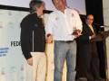 Presentación en un evento de la Federación Mundial de Vela. Un hombre recibe un premio mientras otro lo felicita. En el fondo, una pantalla con logotipos de la organización.