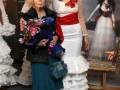 Dos mujeres vestidas con trajes de flamenca posan frente a un fondo con la imagen de una mujer vestida de blanco y rojo. La escena parece estar en un evento o exposición relacionado con la moda flamenca, posiblemente una feria de modas o una exposición de vestidos tradicionales.