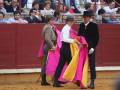 Los toreros sostienen capotes de colores brillantes mientras preparan la corrida. El ambiente es animado con espectadores en el fondo.