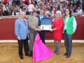 Un grupo de personas, incluyendo una mujer con un vestido rosa y botas altas, sostienen un premio junto a otros individuos en una arena de toros. La escena parece ser parte de un evento deportivo o cultural, con una multitud observando desde la tribuna.