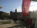 Callejón con edificio histórico y bandera roja en primer plano, con gente caminando alrededor.