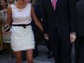 Una mujer viste un vestido blanco con detalles negros y lleva una bolsa de mano. Un hombre en traje negro mira hacia ella, ambos parecen estar en un evento formal.