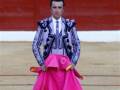 Un torero en traje tradicional con capa rosa y pañuelo de color púrpura, en un escenario de toros con fondo rojo.