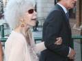 A una mujer con cabello blanco y gafas de sol, posiblemente la actriz Carmen Dell'Orefice, caminando con un hombre en traje. La escena parece ser una celebración o evento público, posiblemente en Italia, dada la arquitectura y el ambiente.