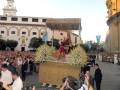 Una procesión religiosa en una ciudad histórica, con un carro decorado y flores en el centro. La gente observa la procesión mientras una cámara de televisión filma el evento. En el fondo, edificios con detalles arquitectónicos y banderas ondean en la plaza.
