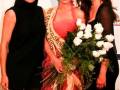 Tres mujeres posan juntas, una con un vestido rojo y corona, otra con un vestido negro y la tercera con un vestido rojo. La imagen parece ser de una competencia o evento de belleza.