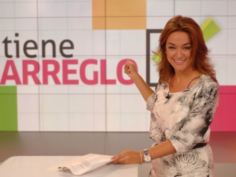 Una mujer sonríe y levanta el puño en un estudio de televisión, con una pantalla que dice "tiene ARREGLO" y colores vivos al fondo.