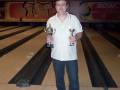 Un hombre sostiene dos trofeos en una pista de bowling, con luces iluminando el espacio.