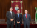 Tres hombres en trajes posan frente a una cortina roja con un escudo y un escudo de armas en el centro. La imagen parece ser de una reunión oficial o ceremonia formal.
