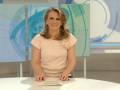 Una presentadora de noticias en un estudio con un fondo azul claro y una mesa blanca en el frente.