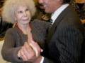 Una mujer con cabello blanco y una persona en traje sonríen mientras se tocan las manos. La imagen parece ser de un evento formal o ceremonial.