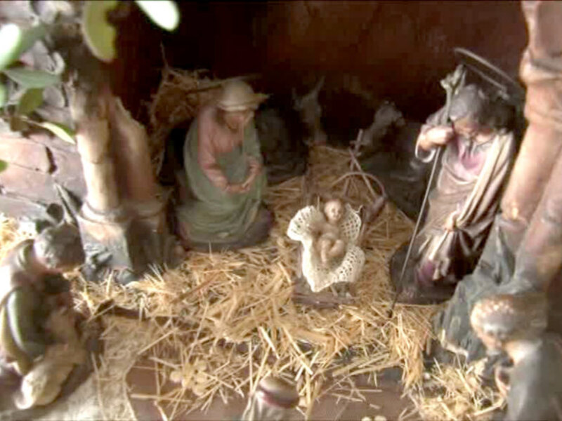 Escena navideña con un bebé Jesús en el centro, rodeado de personajes bíblicos. La escena se desarrolla en un establo con paja y madera, iluminada por la luz de una vela.
