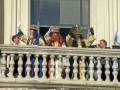 Festividad con personajes en trajes de lujo saludando desde un balcón histórico.