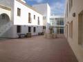 Un pequeño patio con una fuente en el centro, rodeado por edificios blancos y azules. Hay dos bancos para sentarse y una escalera de acceso a las ventanas del edificio. El cielo está despejado y el suelo es de baldosas rojas.