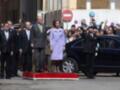 A una mujer en un abrigo largo y azul, posiblemente la reina Letizia de España, junto a un hombre en traje formal. La escena parece ser durante una ceremonia o desfile, con un coche negro en el fondo y personas observando.