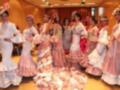 Una escena festiva con mujeres vestidas en trajes tradicionales de flamenca, posando y bailando. La imagen muestra detalles como falda larga con volantes y corona de flores en la cabeza. El ambiente parece ser un evento cultural o competencia de baile flamenco.