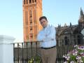 En el fondo, se puede ver la Giralda de Sevilla, una torre medieval icónica. En primer plano, un hombre con camisa blanca y pantalones beige posa en una terraza con flores.