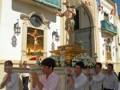Una procesión religiosa con personas vestidas de blanco y rojo, llevando una cruz dorada. Detalle de la imagen: estatuas religiosas en un altar, banderas colgando y una iglesia al fondo.