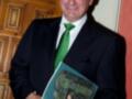 Un hombre vestido con un traje negro y corbata verde sosteniendo un libro titulado "Heliocéfalo". La imagen se centra en su rostro y torso, destacando la expresión serio del hombre.