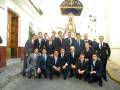 Un grupo de hombres en trajes formales posa frente a una imagen de la Virgen, posiblemente para un evento religioso o cultural en España.