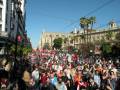Una multitud de personas camina por una calle con edificios históricos y árboles en el fondo. La imagen muestra un ambiente festivo o de protesta, con banderas rojas y pancartas en la mano izquierda. El cielo es claro y el clima parece ser cálido.