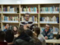 Un hombre con una camiseta de rayas y gafas está en el centro de la imagen, hablando a un grupo de personas sentadas frente a él. La escena se desarrolla en una biblioteca con estanterías llenas de libros.