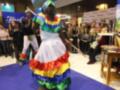 Una mujer en un traje de colores vibrantes realiza una danza tradicional, con otros participantes observando. La imagen se toma en un evento o feria, con stands y publicidad al fondo.