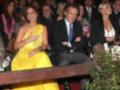 Una mujer en un vestido amarillo brillante se sienta en el público, rodeada de otros asistentes. La escena parece ser una gala o evento formal con vestimentas elegantes y un ambiente sofisticado.