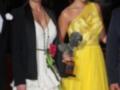 Dos mujeres posan con un premio en una alfombra roja. Una lleva un traje blanco y la otra, un vestido amarillo con una falda plisada. La mujer de la derecha sostiene un trofeo con un águila en su portada.