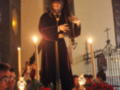 Imagen de una estatua religiosa en un altar con velas encendidas y flores rojas, rodeada por personas observando la escena.