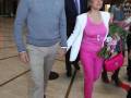 Una pareja camina juntos en un interior moderno, ella con traje rosa y flores, él en suéter gris y pantalones beige.
