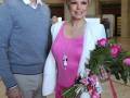 Una pareja posa con un hombre en suéter azul y una mujer con traje blanco y pantalones rosa brillante, sosteniendo un ramo de flores.