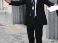 Un hombre en traje negro con una medalla colgada, extendiendo sus brazos hacia el frente mientras sostiene un papel en la mano izquierda. El fondo muestra una calle con una estructura de hormigón y un edificio en el lado izquierdo.