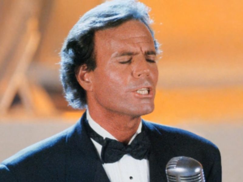 El cantante, con cabello gris y un traje formal, está en el escenario, posiblemente durante una ceremonia de premios. Se puede ver un micrófono en su frente, lo que sugiere que está cantando o hablando. La iluminación y el fondo suave indican un ambiente formal, típico de eventos de reconocimiento.