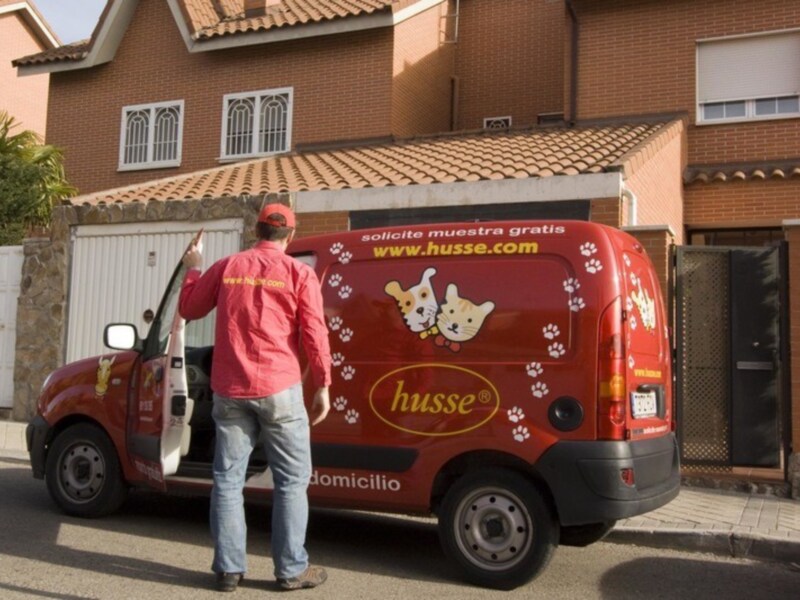 Un hombre en una camiseta roja y un gorro de béisbol, con una camioneta roja que dice "husse" y "domicilio". La imagen muestra una casa con un toldo rojo, ventanas con persianas y una puerta de metal.