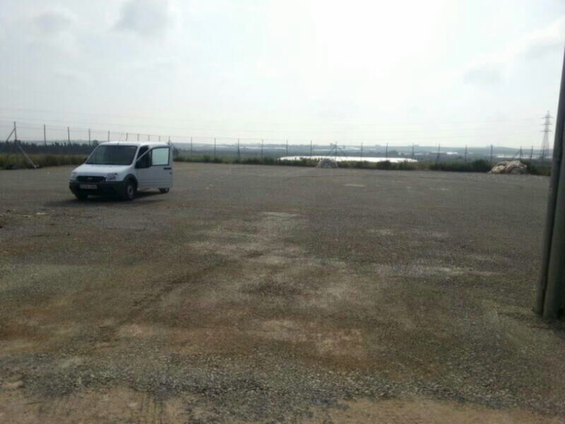Una camioneta blanca está estacionada en un área desierta con una cerca metálica al fondo. La superficie es de tierra seca y despejada, con algunas áreas cubiertas de hierba baja. El cielo está nublado y la atmósfera parece tranquila.