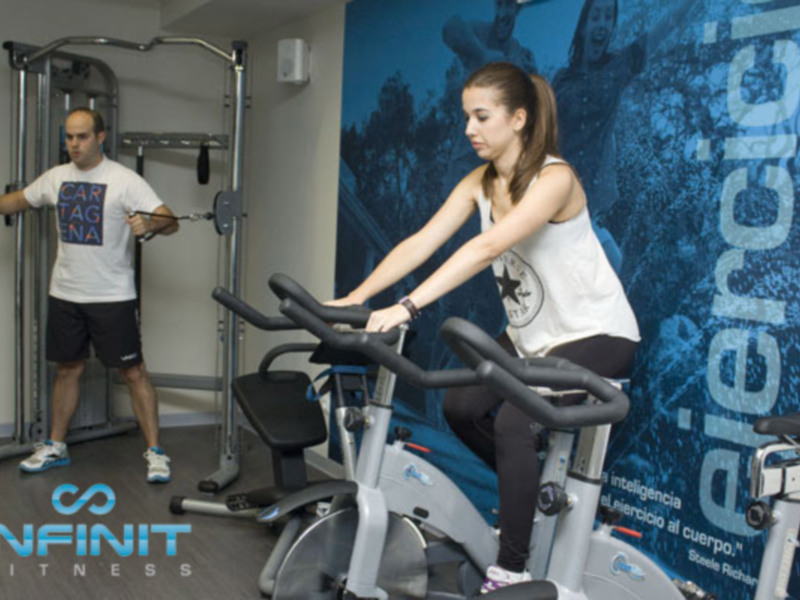 En una sala de fitness, un hombre y una mujer se ejercitan. La mujer está en una bicicleta estática, mientras que el hombre maneja un equipo de musculación. La pared detrás de ellos tiene una imagen de árboles y la palabra "Ejercicio" en letras grandes. En el suelo, se puede ver un logo de Infinit Fitness.