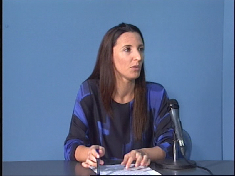 Una mujer con cabello largo y oscuro, vestida con una camiseta de manga larga azul y negra, se encuentra sentada en un escritorio frente a una pared azul. Está escribiendo algo en un papel con una pluma y mirando hacia la cámara, mientras que un micrófono está colocado a su lado.