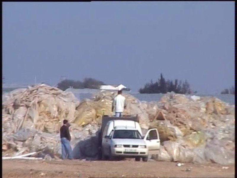 Dos hombres observan una pileta de basura en la playa, con un vehículo blanco estacionado cerca. La escena se ve tranquila y desolada, con un cielo claro en el fondo.