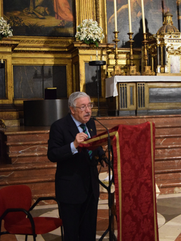 En una iglesia llena de luces doradas y adornos, un hombre en traje negro se encuentra frente a una tribuna con mantel rojo. Detrás de él, un altar con velas y pinturas antiguas ilumina la atmósfera serena. La imagen captura un momento de reflexión y solemnidad en un espacio sagrado.