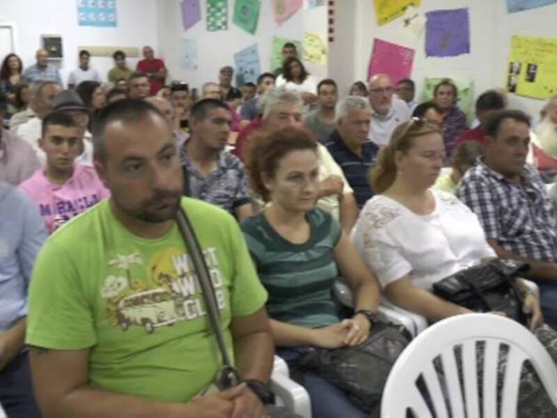 Un grupo de personas asistentes a una reunión o conferencia, sentados en sillas blancas. La sala está decorada con carteles de colores y un hombre en el centro lleva una mochila.