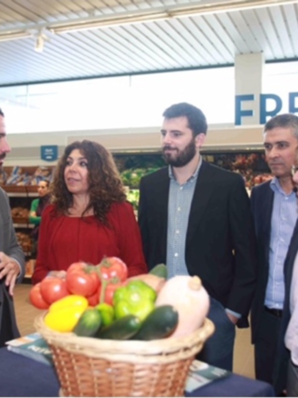 En un supermercado moderno, cuatro personas conversan amistosamente frente a una cesta llena de frutas y verduras frescas. La atmósfera es relajada, reflejando la importancia de la alimentación saludable y la sostenibilidad en el comercio.