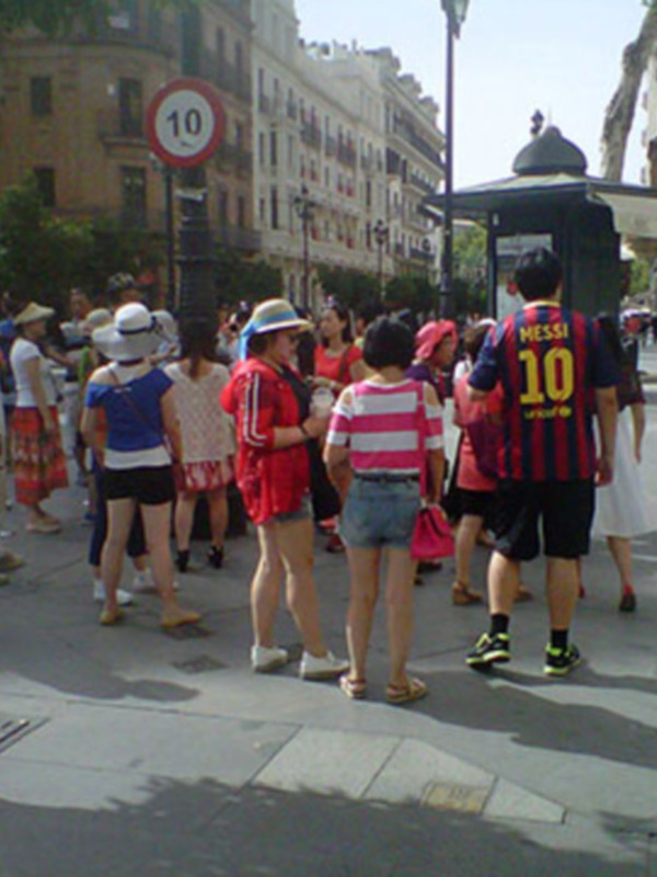 Se observa a varias personas caminando en una calle de ciudad, con edificios al fondo y un poste indicador de velocidad. La escena parece ser un día normal en un entorno urbano con gente caminando y algunas personas paradas, posiblemente esperando en una parada de autobús.