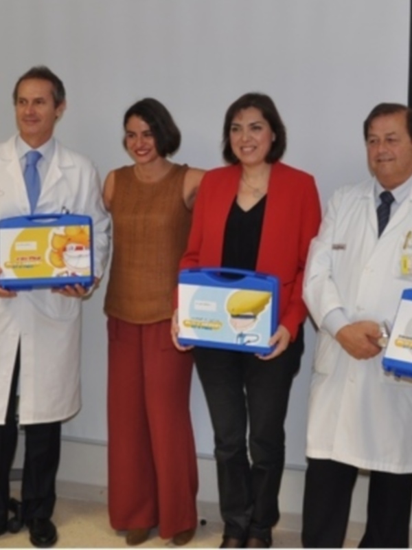 Un grupo de personas, incluyendo médicos y personajes de dibujos animados, sostienen cajas con el logo de un hospital. La escena parece ser una presentación o evento médico, posiblemente relacionado con la prevención de enfermedades infantiles.