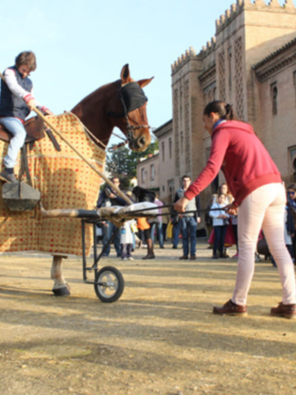 Una persona monta un caballo de juguete grande, mientras otra camina a su lado. El fondo muestra una arquitectura histórica y un grupo de espectadores observando la escena.