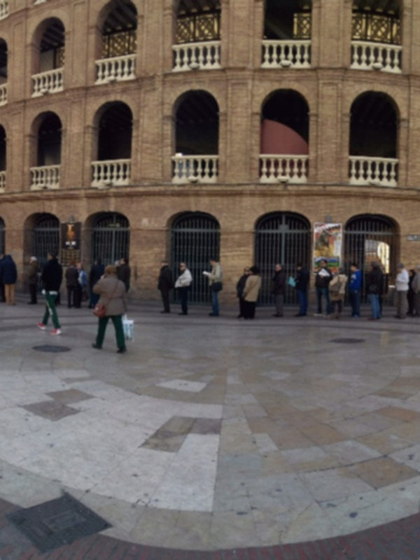 Se puede ver una plaza con un edificio de ladrillo rojo en el fondo. La plaza está decorada con una forma geométrica en el suelo y hay muchas personas de pie, algunas esperando. El edificio tiene arcos y balcones.
