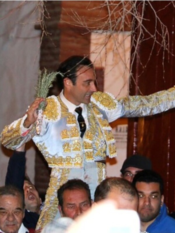 Un torero vestido con un traje tradicional de color dorado y blanco, con una saya y capote, salta sobre un grupo de personas. La escena parece ser en un evento cultural o festivo, con una pared roja y árboles en el fondo.
